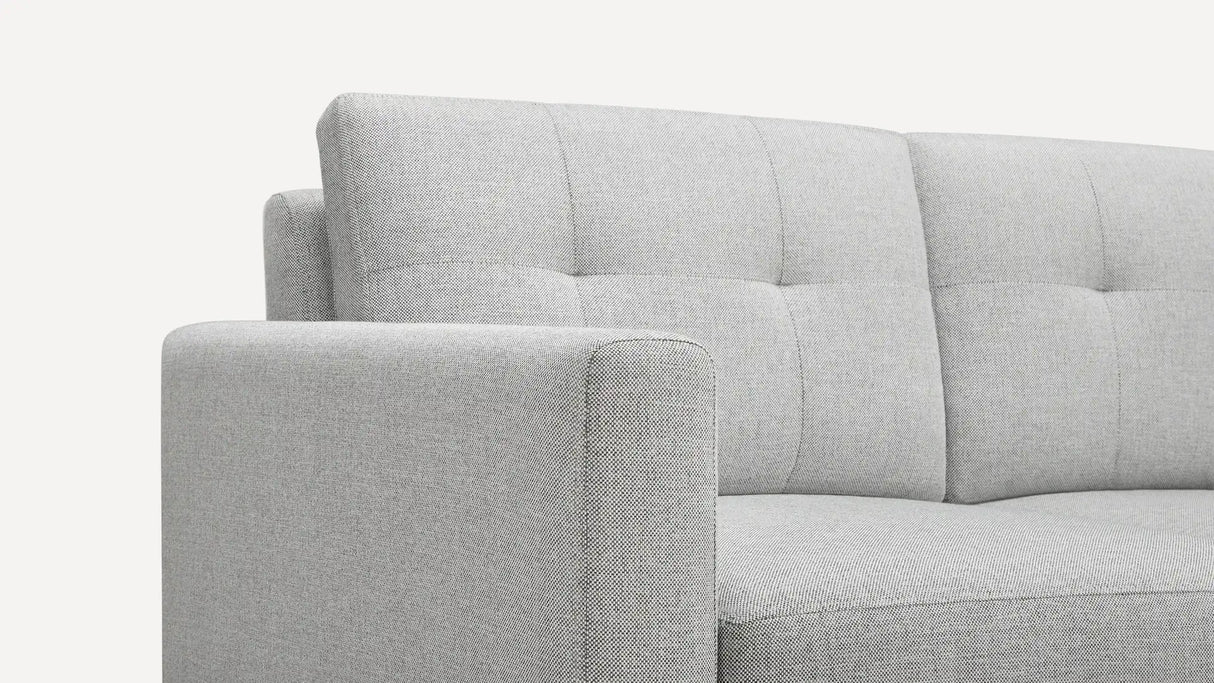 Close-up of the Nomad Fabric Sofa in light gray, showcasing its tufted backrest and sleek, modern lines. The fabric texture is clear, with the image highlighting the armrest and part of the backrest.
