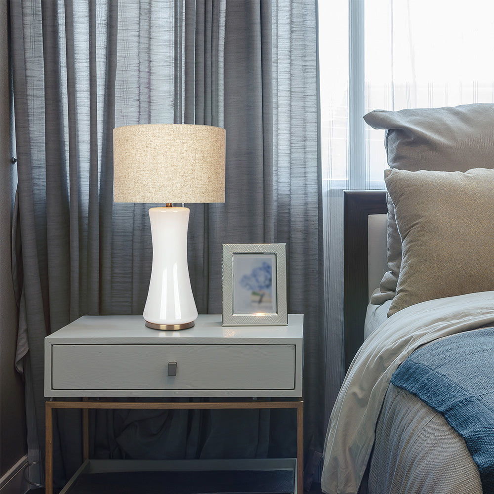 A bedside scene with a modern Cascade Lamp in beige/gold/white, a small framed photo, and a gray nightstand. Behind are gray curtains partially covering a window beside a neatly made bed featuring a beige headboard and layered pillows.