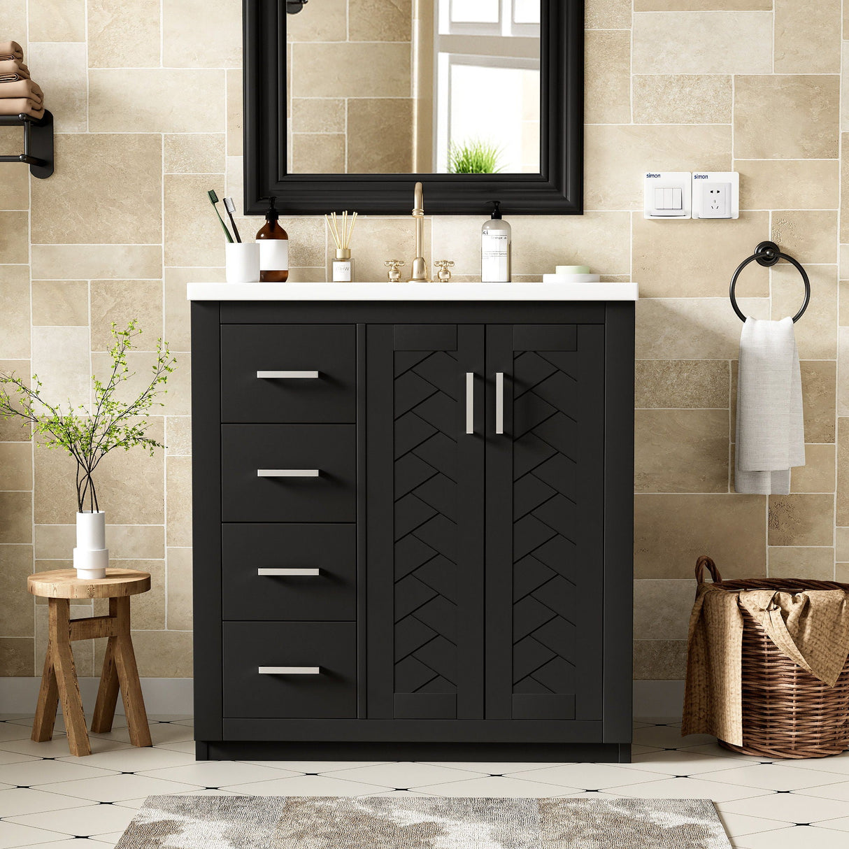 A modern bathroom with beige tile walls features a dark freestanding vanity set including a ceramic sink, three drawers, and soft closing doors. A large mirror hangs above. A wooden stool with a plant and a woven basket are nearby. A hand towel is beside the sink.