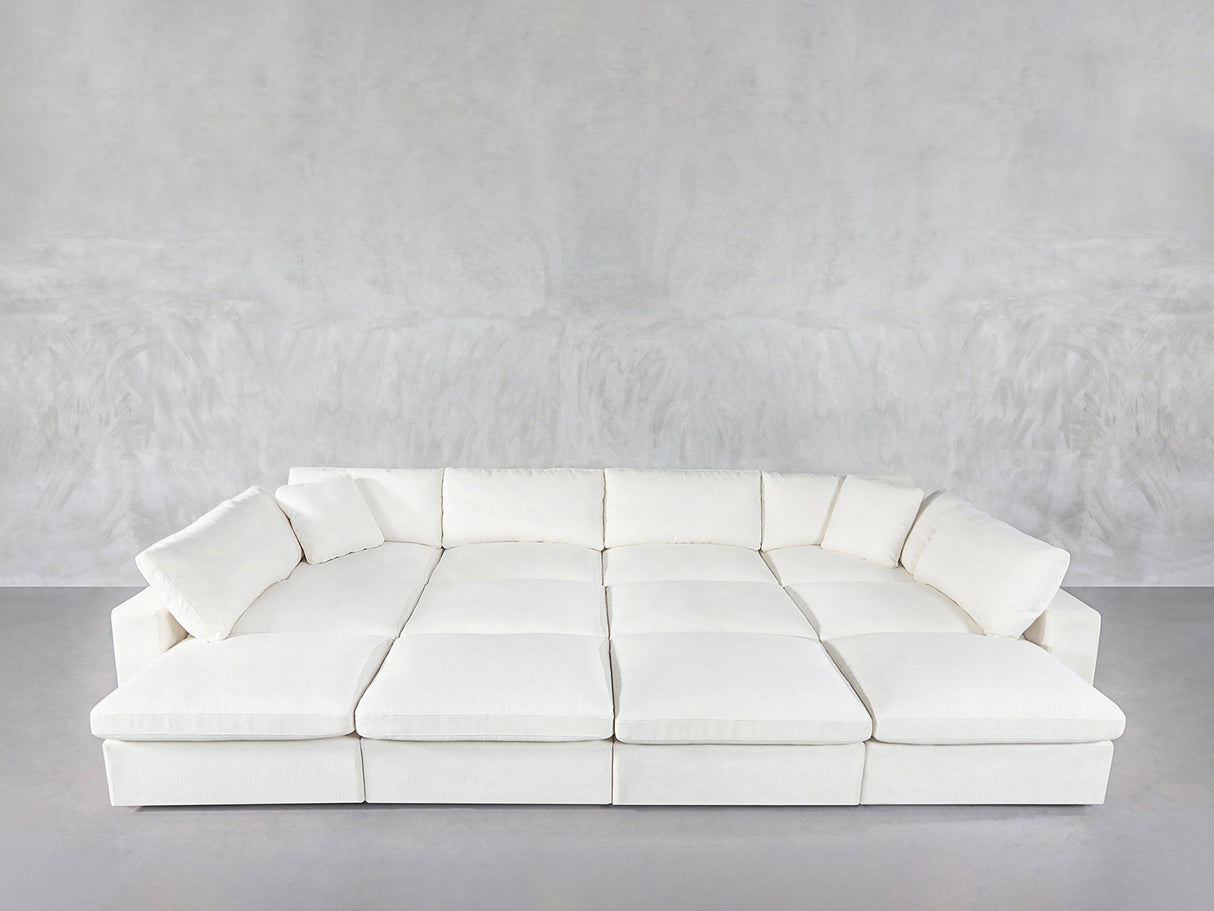 A spacious 12-Seat Deep Modular Daybed with plush cushions and wide seats sits against a light gray textured wall in a minimalist room featuring a concrete floor.