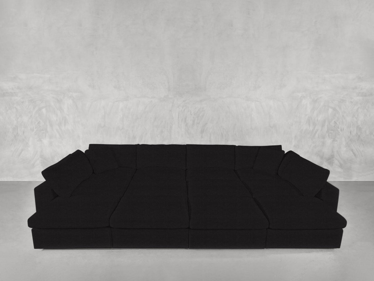 A 12-Seat Deep Modular Daybed in black, featuring multiple cushions, is set against a light gray textured wall on a smooth, light-colored floor.