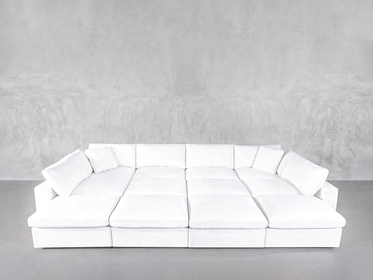 A 12-Seat Deep Modular Daybed with white upholstery and multiple cushions sits against a plain light gray textured wall on a gray floor.