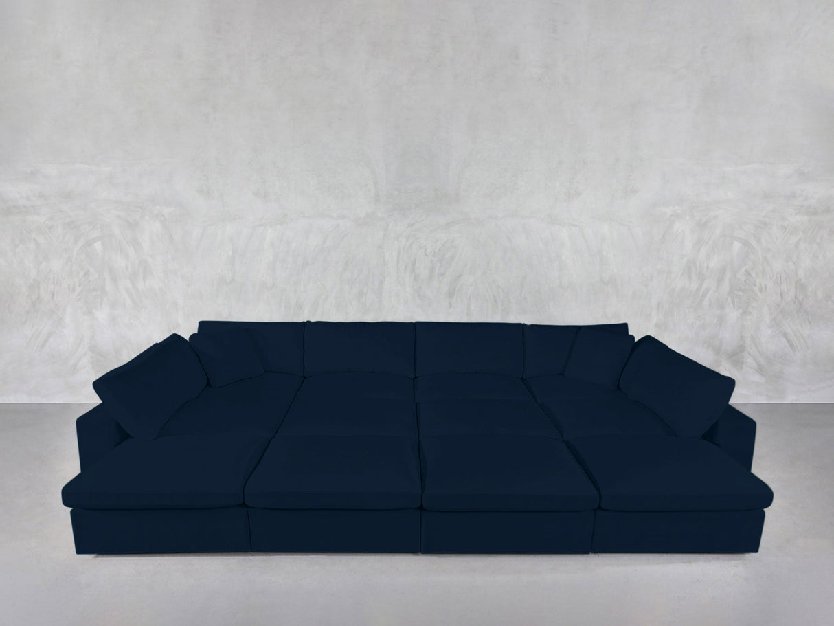 The 12-Seat Deep Modular Daybed, a spacious and modern dark blue sectional with wide seats and multiple cushions, is placed against a simple light gray textured wall and floor.