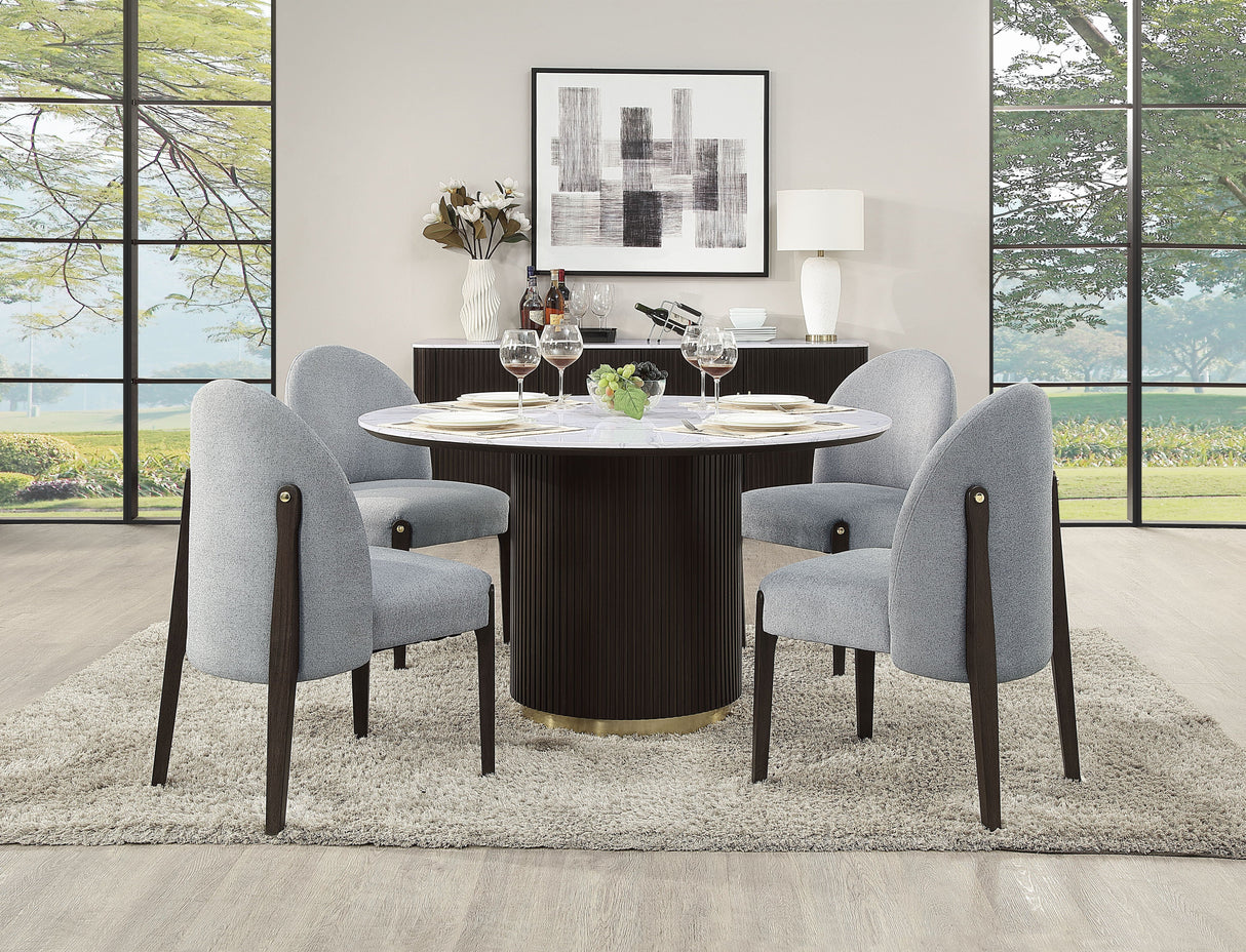 A modern dining room features the Clayten Around Dining Table in Marble/Espresso, set with plates and wine glasses, surrounded by four upholstered chairs. Neutral-toned walls and floors enhance large windows revealing outdoor greenery, abstract wall art, and a decor-adorned sideboard.