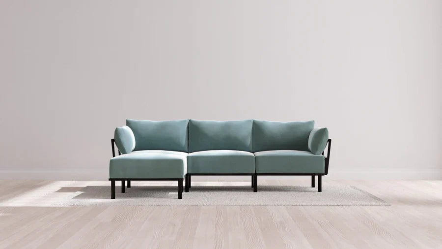 A modern light blue sectional sofa with black legs is placed on a light gray rug. The background features a plain light gray wall and a light wooden floor. The setting is minimalist and spacious.