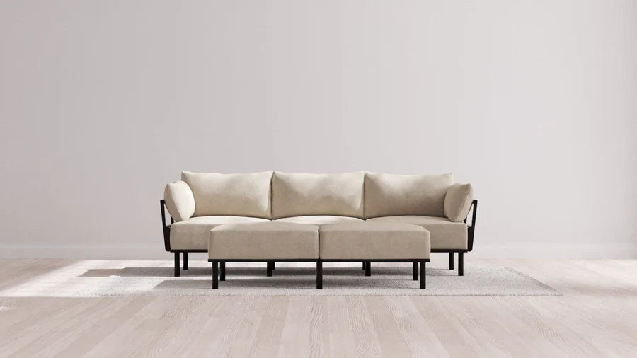 A modern, minimalist living room featuring the Indoor Jack 3-Seater Sofa + Bench Ottoman in Balsa set on a light grey rug. The walls are plain white, and the sunlight casts soft shadows on the wooden floor.
