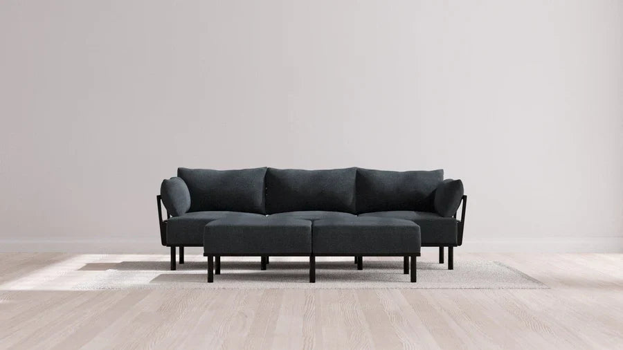 A contemporary sectional sofa in Balsa, complemented by three cushions, is set on a light gray area rug within a minimalist room featuring light wooden floors and plain off-white walls.