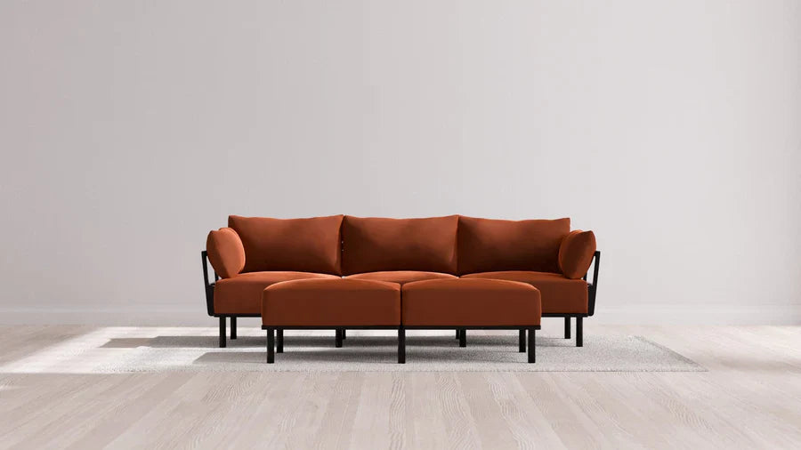 A modern living room features the Indoor Jack 3-Seater Sofa and Bench Ottoman in Balsa, showcasing a vibrant orange upholstery with black legs and armrests. The stylish set is elegantly placed on a light gray rug, framed by a plain white wall and wooden flooring.
