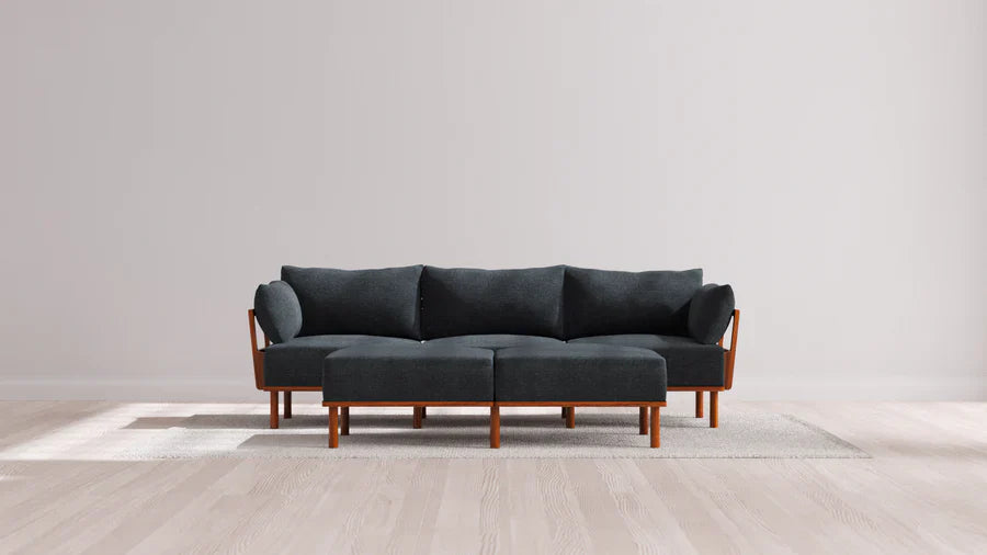 A modern Indoor Jack 3-Seater Sofa with Bench Ottoman in Balsa, featuring wooden legs and a dark gray tone, is placed on a light-colored rug in a minimalist room. The floor is made of light wood, and the walls are painted a neutral shade, creating a clean and spacious ambiance.