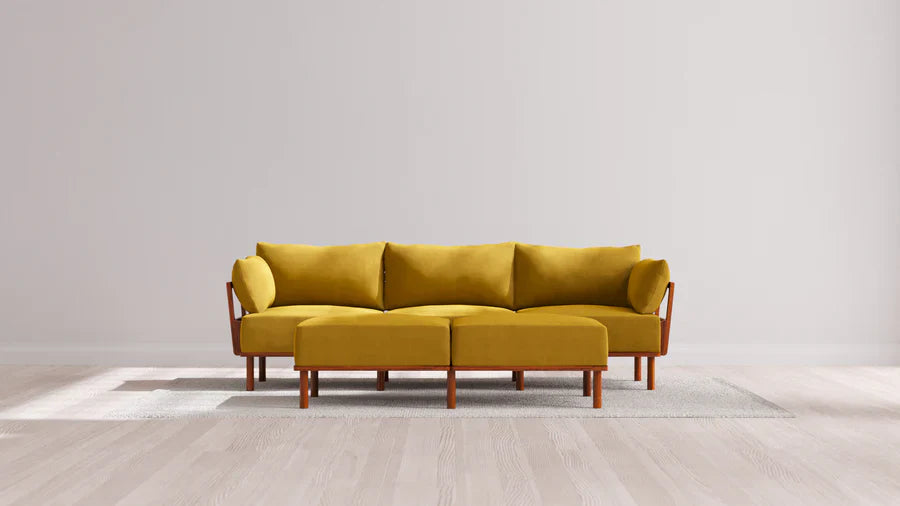 The Indoor Jack 3-Seater Sofa + Bench Ottoman in Balsa, with its mustard yellow hue and wooden legs, is placed atop a light gray rug in a minimalist room characterized by a light wood floor and plain off-white walls. The sofa showcases clean lines and comes with two matching bolster pillows.