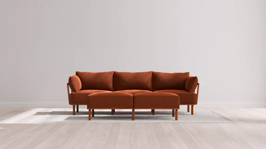 In a contemporary living room, an Indoor Jack 3-Seater Sofa with a Bench Ottoman in Balsa is elegantly placed on a light gray rug over a wooden floor, set against a plain white wall.