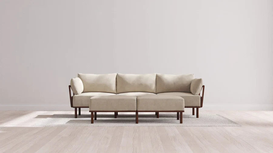 An Indoor Jack 3-Seater Sofa with a Bench Ottoman in Balsa is placed on a light-colored rug in a bright room featuring wooden flooring and plain walls. The sofa's wooden arms and legs enhance the neutral, modern aesthetic.