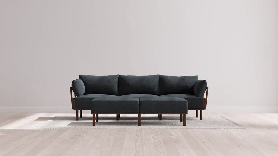 The Indoor Jack 3-Seater Sofa + Bench Ottoman in Balsa, featuring a modern dark gray color with wooden legs and armrests, is elegantly placed on a light-colored rug in a minimalist room with pale wooden floors and a plain white wall. This sofa boasts comfortable cushions and a sleek, contemporary design.