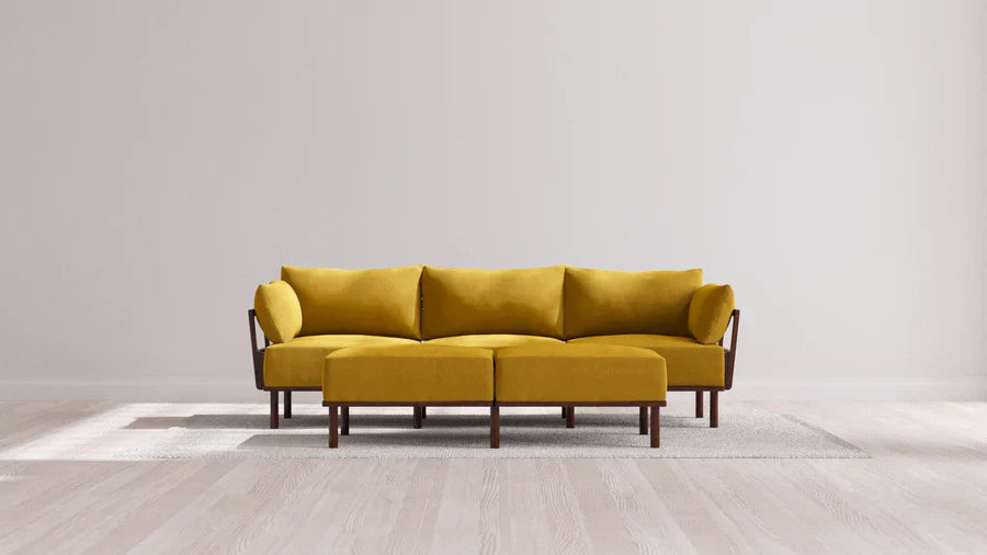A modern living room featuring the Indoor Jack 3-Seater Sofa and Bench Ottoman in Balsa, set atop a light gray area rug. The space is enhanced by light wood flooring and plain white walls, contributing to a minimalist and spacious atmosphere.