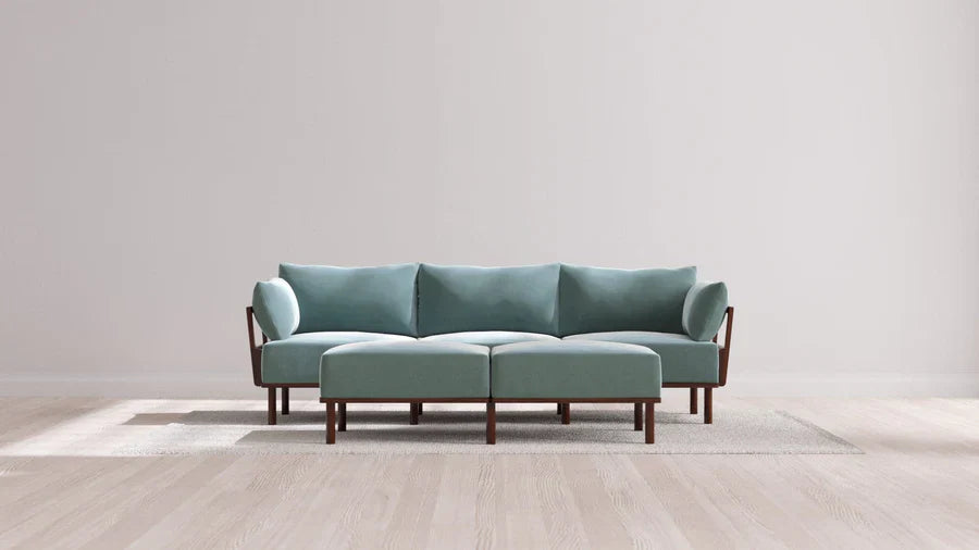 A contemporary living room showcases the Indoor Jack 3-Seater Sofa along with a coordinating Bench Ottoman, both in a soft balsa hue. These pieces feature elegant wooden legs and armrests, resting on a light area rug. The room's decor is minimalist and neutral, boasting a wooden floor and a simple white wall in the background.