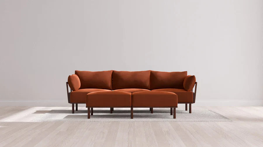 A modern, minimalist living room showcases the Indoor Jack 3-Seater Sofa and matching Bench Ottoman in Balsa, their rust color beautifully complemented by wooden legs. These pieces rest on a light gray rug atop light wooden flooring, against a backdrop of plain, neutral-toned walls.