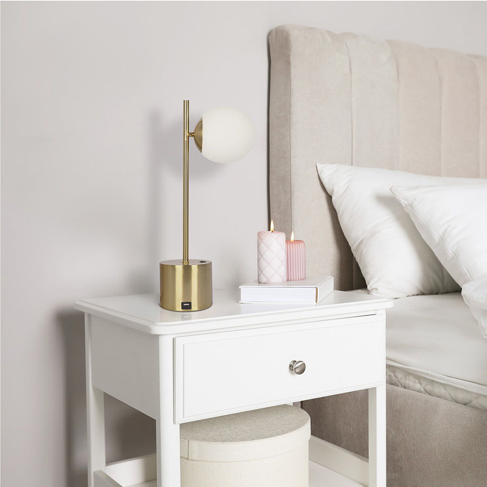 A modern bedroom showcases a white nightstand with the "Ethereal" gold table lamp, two pink candles, and a book. The beige upholstered bed and white pillows in the background enhance the clean and minimalist aesthetic.