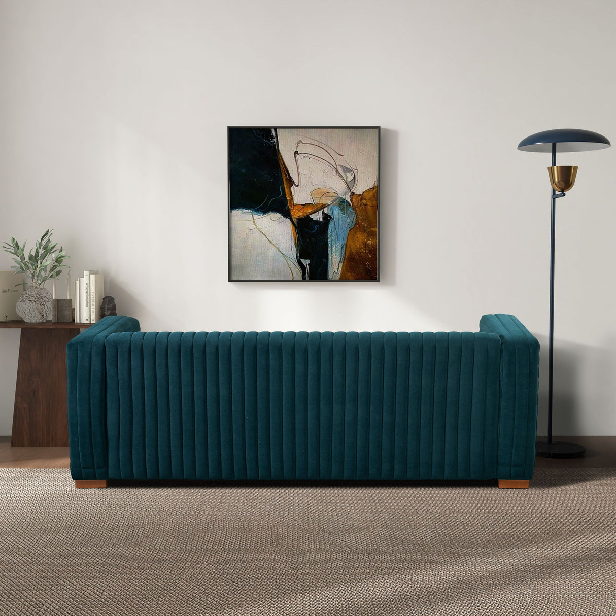 A living room featuring the Elara - Upholstered Sofa in teal, backed by an abstract painting. A modern floor lamp stands on the right, while a wooden table with plants and books sits to the left. The carpeted floor adds warmth to the scene.