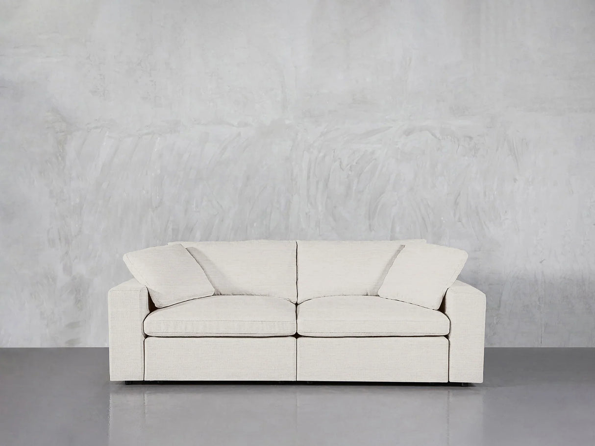 A modern, white 2-Seat Modular Loveseat with seat and back cushions is set against a plain gray, textured wall. The smooth grey floor enhances the minimalist and clean aesthetic.