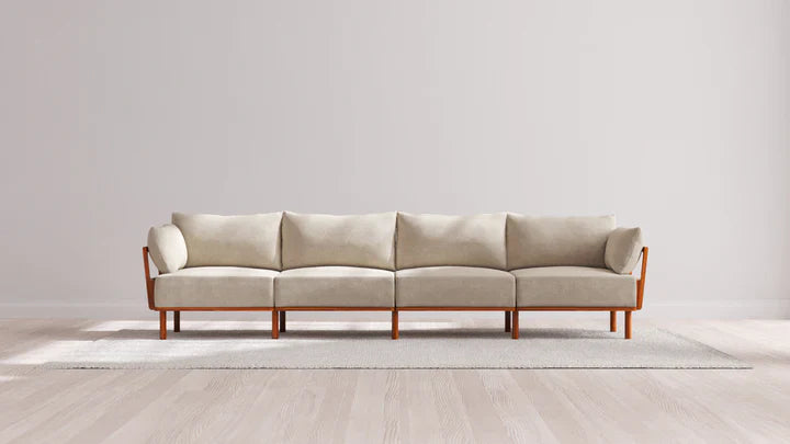An Indoor Jack 4-Seater Sofa in Balsa, featuring a wooden frame and armrests, sits on a light gray rug in a minimalistic room with light wood flooring and a plain white wall.