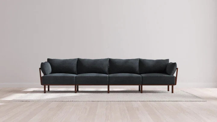 The Indoor Jack 4-Seater Sofa in Balsa, featuring a sleek, modern design with its dark gray upholstery and wooden frame and armrests, is positioned on a light rug. The backdrop consists of a smooth, light gray wall paired with a wooden floor, establishing a minimalist and spacious interior ambiance.