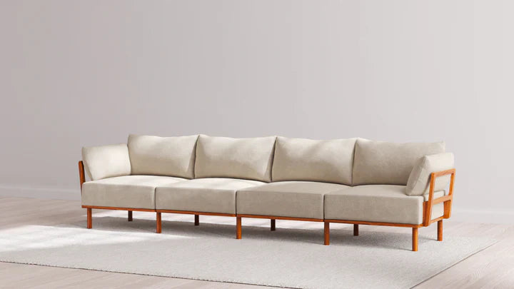 An Indoor Jack 4-Seater Sofa in Balsa, featuring a modern design and wooden frame, sits on a light gray rug. The background showcases a plain light-colored wall and hardwood flooring.