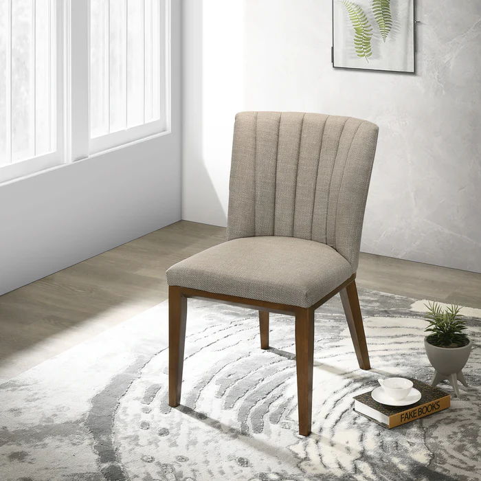 An Elmwood Stylish Dining Chair, beige and wooden-framed, sits on a circular patterned rug in a bright room. Sunlight pours through large windows, lighting up a book and potted plants nearby.