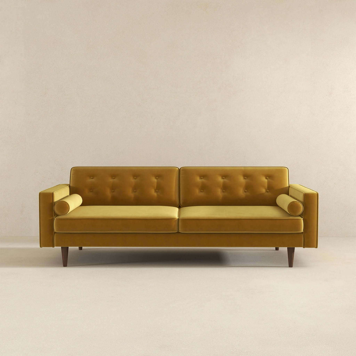 Against a beige wall stands the Casey mid-century modern sofa in mustard yellow, featuring a tufted backrest, cylindrical arm pillows, wooden legs, and a minimalist design.