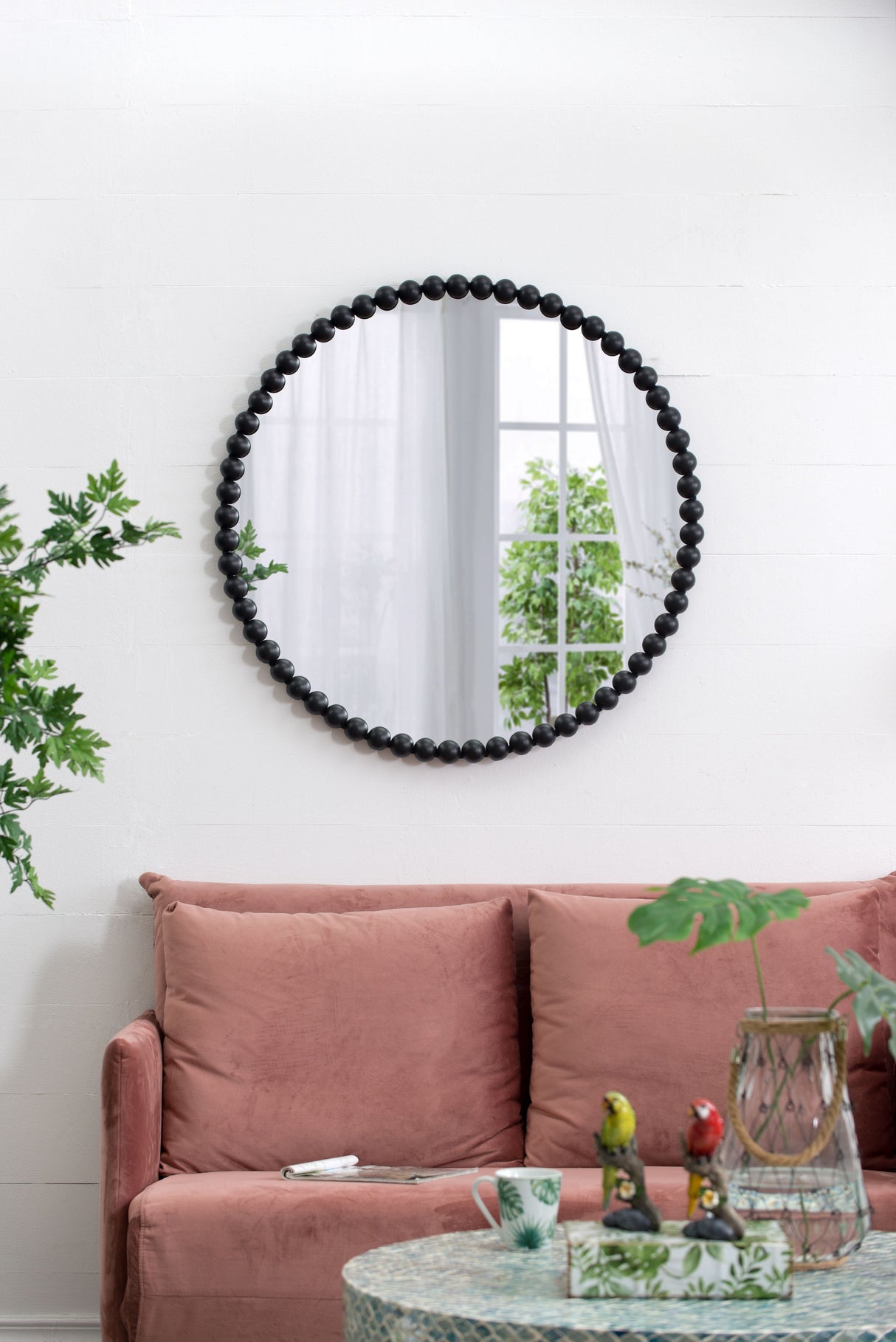 A Circle Mirror with Metal Beaded Frame in black adorns a white wall, reflecting a window with white curtains and outdoor greenery. Beneath it sits a pink sofa with assorted pillows and a patterned coffee table featuring decorative figurines and plants.