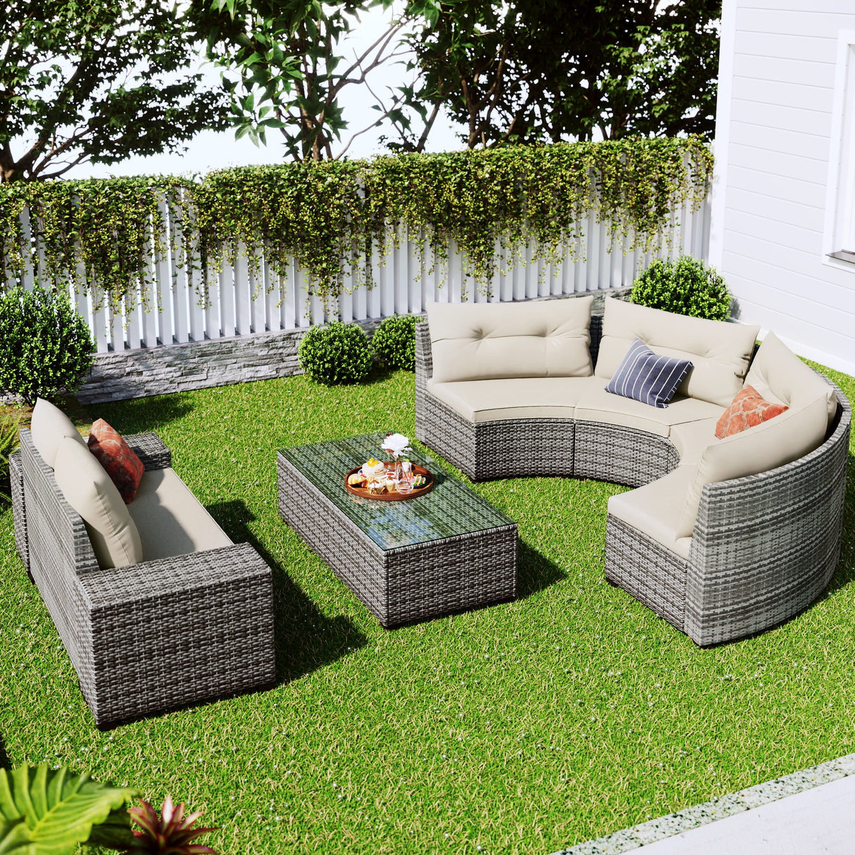 A lush garden features an 8-piece outdoor wicker round sofa set with two curved gray sofas, beige cushions, and a glass-topped rectangular coffee table. Surrounded by a white fence and greenery, this cozy space showcases the all-weather PE rattan set's water-resistant and UV-protected design.