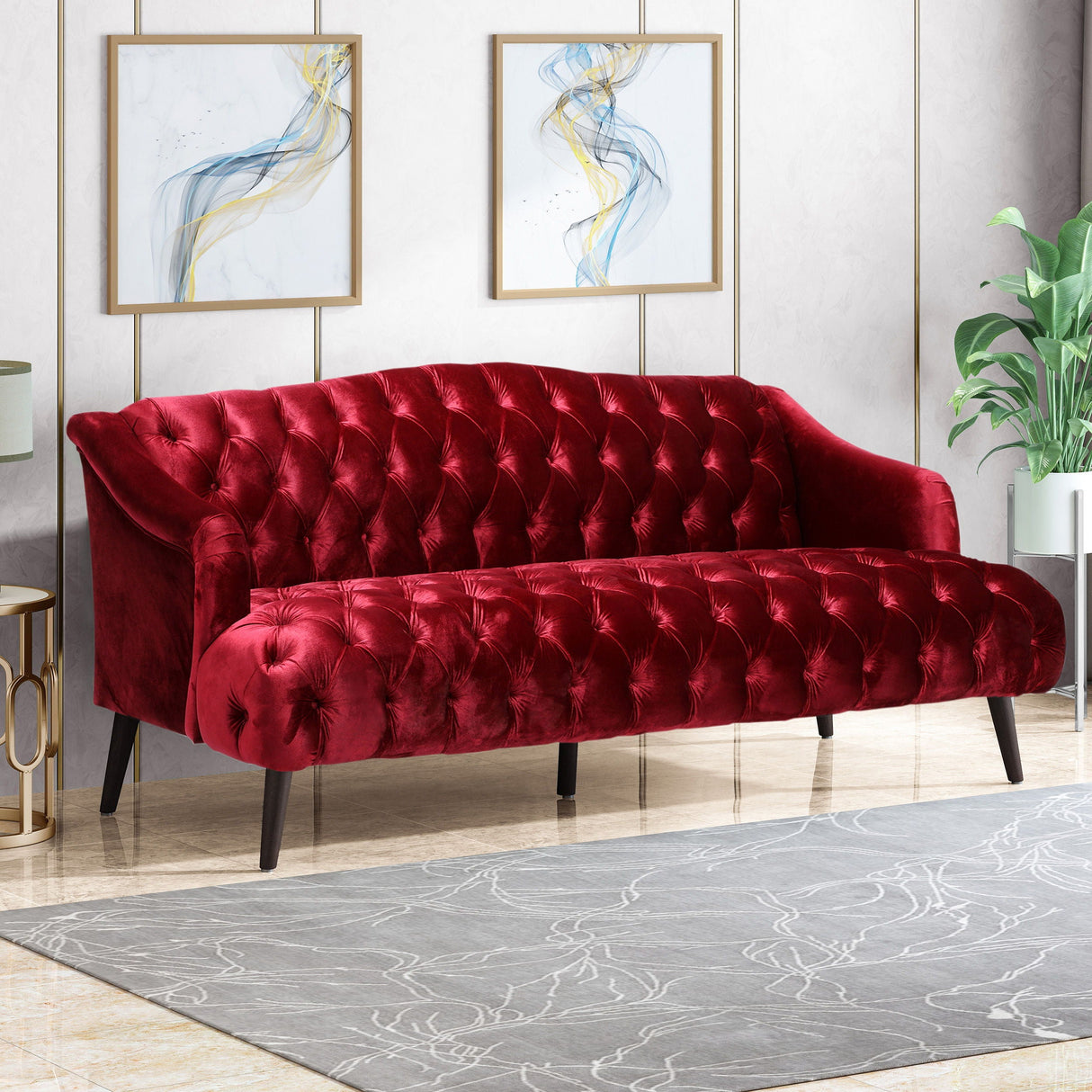 The elegant living room showcases a Comfy 3 Seat Sofa with wooden legs in red velvet and tufted design. The decor includes two abstract paintings, a potted plant to the right, and a patterned gray rug on the floor.