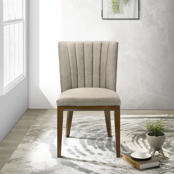 The Elmwood - Stylish Dining Chair with a beige upholstery and wooden legs sits on a patterned gray and white rug. Nearby, a small round table holds books, a plant, and cups. A framed leaf print adorns the textured wall as sunlight streams through a nearby window.