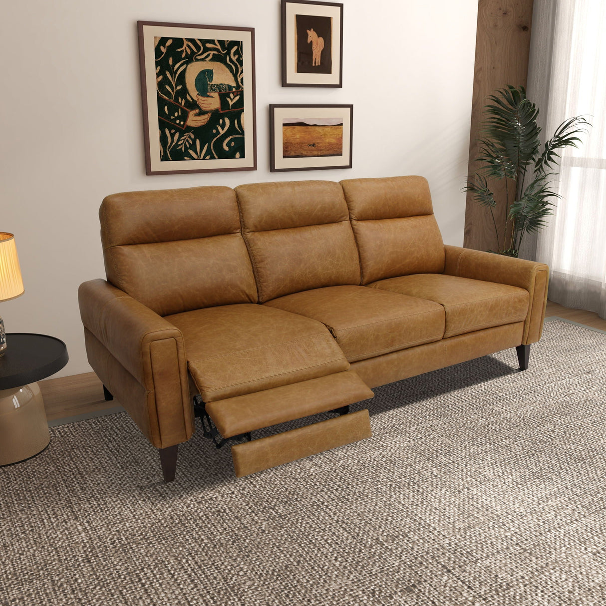 An Elliot Incliner Sofa in brown leather is placed in a living room on a light gray textured rug. Behind it, three framed artworks decorate the wall. A potted plant occupies the corner, while a round side table with a lamp sits to the left.