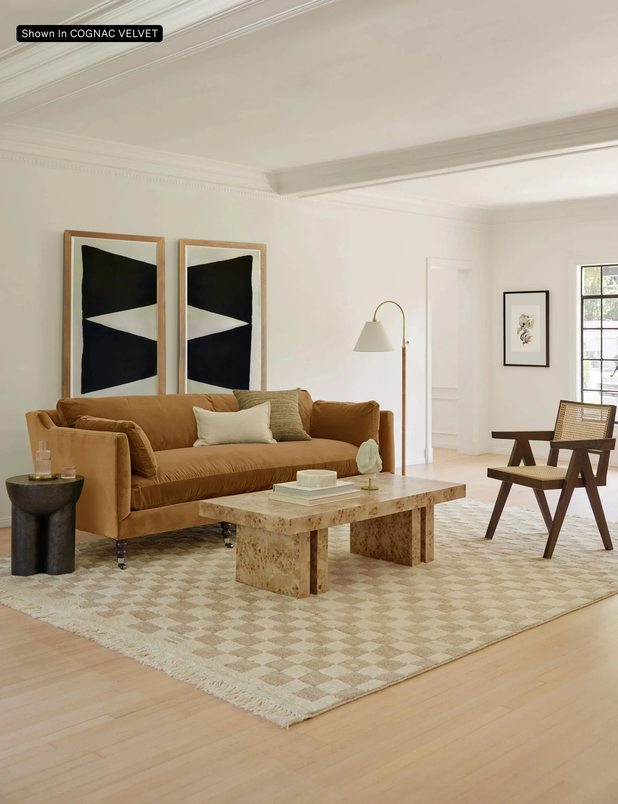 A modern living room showcases the Fabienne Sofa in a rich cognac velvet, complemented by white and black abstract artwork hanging above. A wooden coffee table and chair are set upon a patterned rug, while the space is enhanced with a floor lamp and a side table. The walls and floors feature neutral tones.