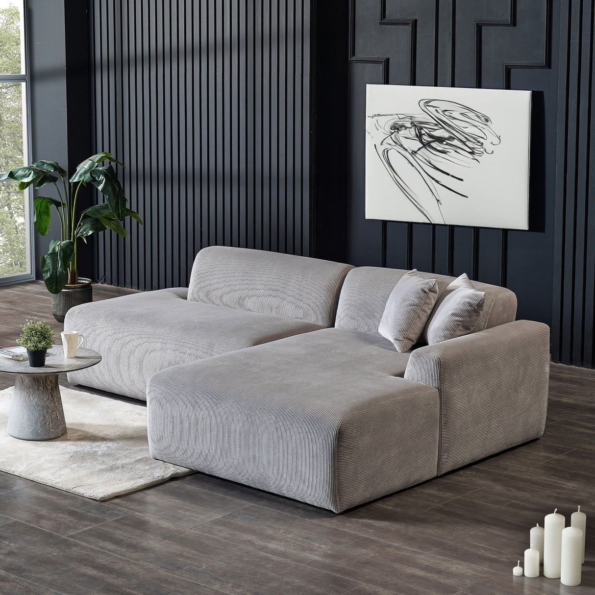 A modern living room showcases the Mar - High-Quality Sectional Sofa in plush gray on a dark wood floor. A round marble coffee table with a potted plant stands before it, abstract art adorns a textured black wall, candles rest nearby, and a large plant sits by the window.
