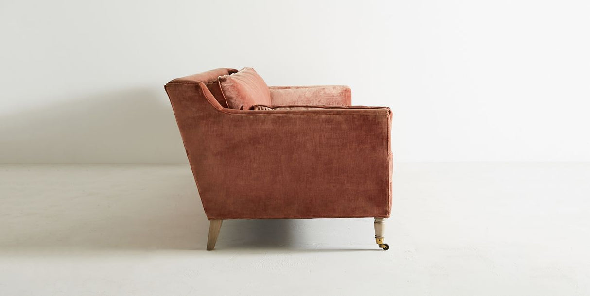 Side view of a pink velvet Leonelle Sofa with plush seat and back cushions. The sofa features slightly tapered wooden legs, with the front right leg on a caster wheel, set against a plain light-colored background.