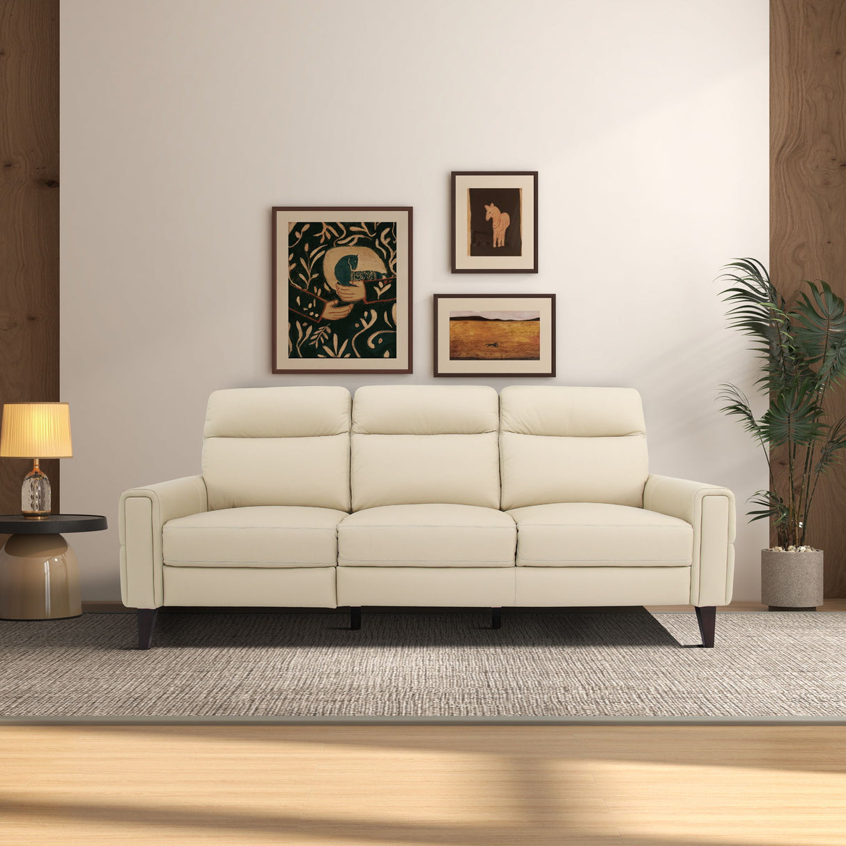 The living room features an Elliot Incliner Sofa Left in cream on a textured rug, with three framed artworks on a light wall. A round table with a lamp is to the left, and a tall plant in a pot is to the right. The space has wooden accents and exudes cozy atmosphere.