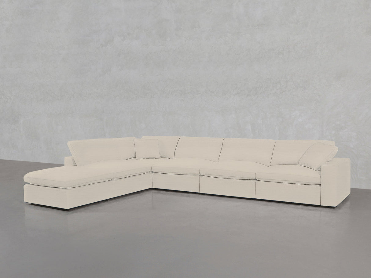The 6-Seat Modular Corner Lounger Sectional features light beige upholstery, wide seat cushions, and pillow-style backrests, with a left-side chaise lounge. It sits on a gray concrete floor against a light gray textured wall.