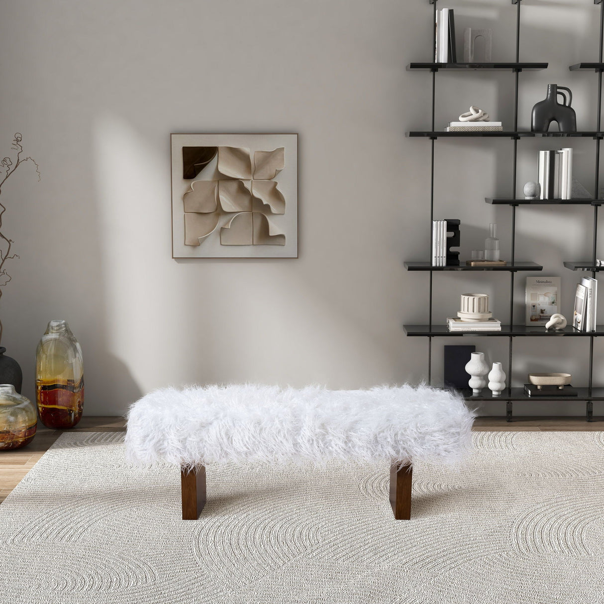 A modern living room showcases the Henley Fur Bench in White Khaki on a textured rug, with a geometric art piece on a light gray wall. A black metal shelf to the right holds decorative items, while a large glass vase rests on the floor to the left.