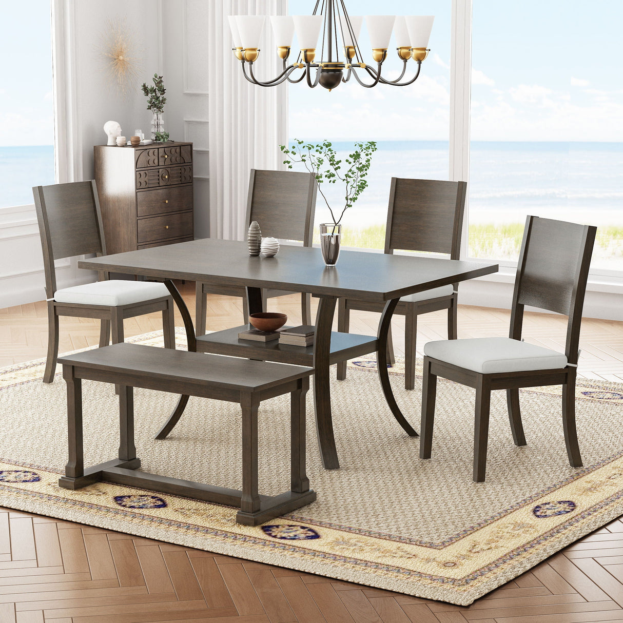 The dining room showcases the Topmax 6-piece wood table set with a bench and four cushioned chairs on a patterned rug. A sideboard is against the windowed wall with a seaside view, and a chandelier hangs above the table, which features a vase with branches.