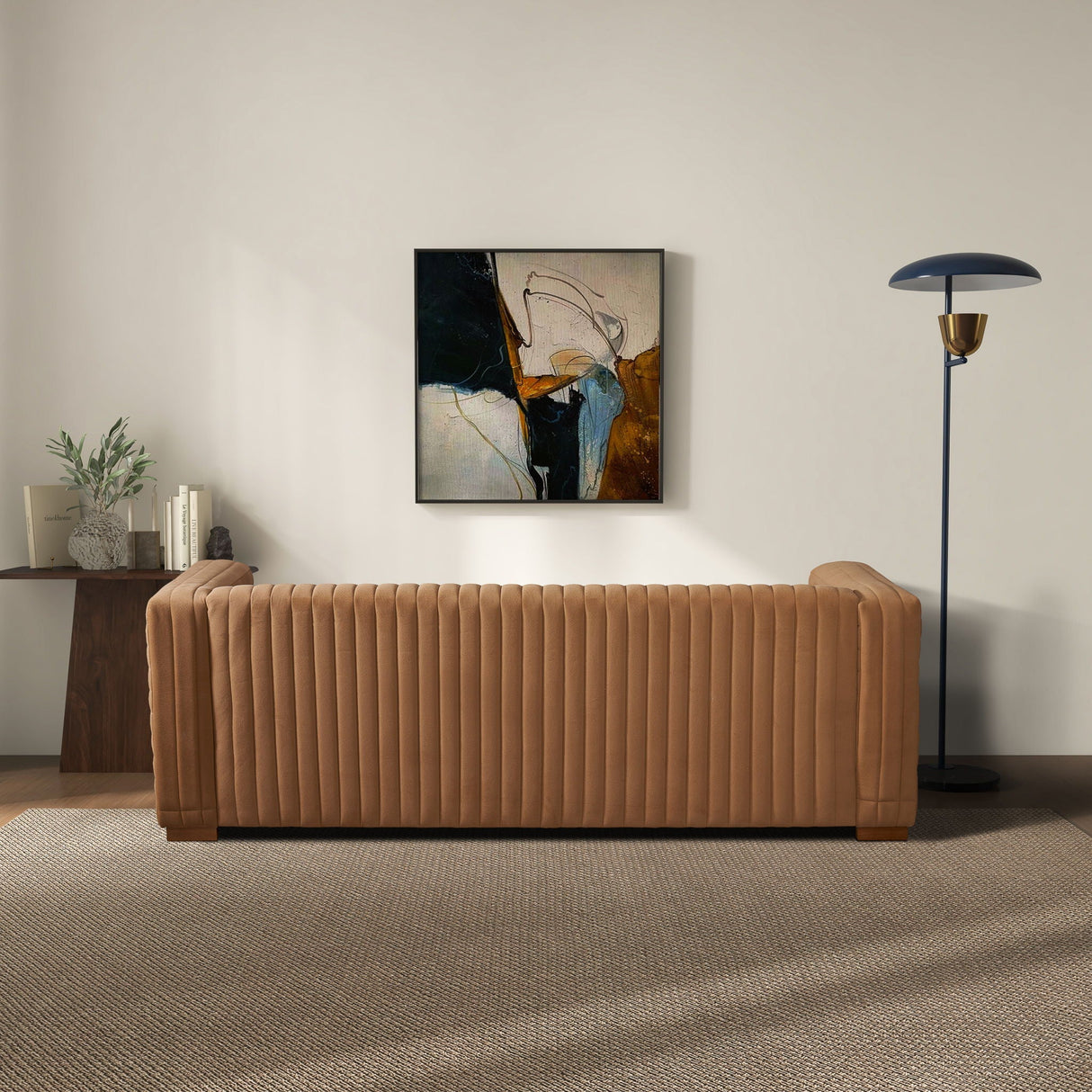 A modern living room features the Elara - Upholstered Sofa, a tall black and gold floor lamp, and a side table adorned with books and a plant. An abstract painting decorates the light-colored wall, while a neutral-toned rug covers the floor.