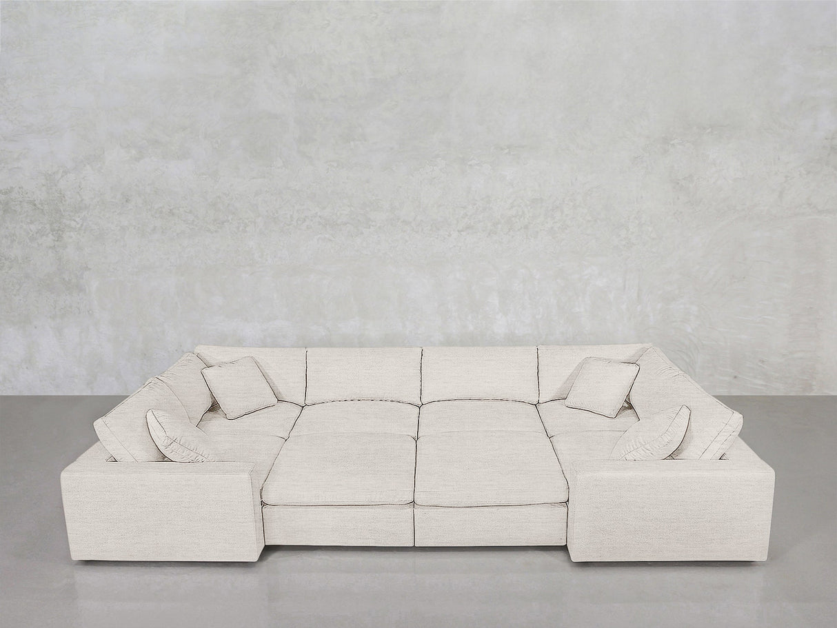 The 8-Seat Modular Closed Pit, a spacious light beige sectional with four matching cushions, is arranged against a plain light grey concrete wall on a smooth grey floor.