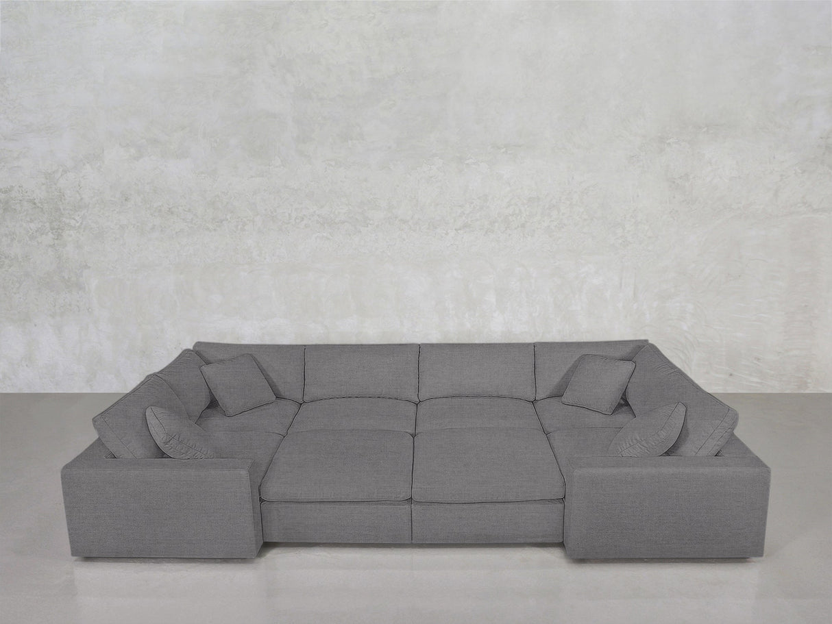 The 8-Seat Modular Closed Pit is a large, modern gray sectional with wide armrests, four matching cushions, and a minimalist design, set against a light gray textured wall and smooth gray floor.