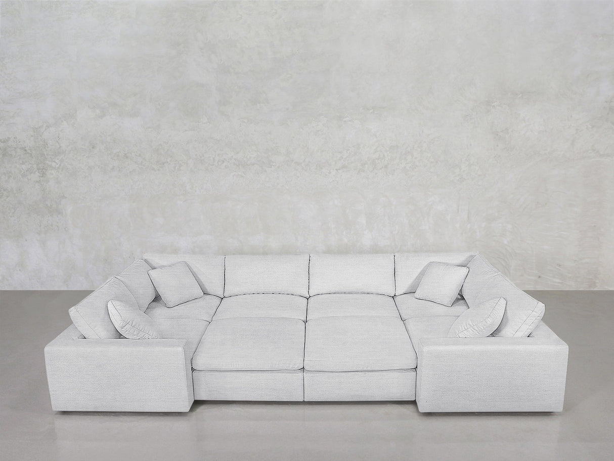 The 8-Seat Modular Closed Pit, a modern light gray sectional with four matching cushions, wide armrests, and a low backrest, is set against a textured light gray wall above a smooth gray floor.