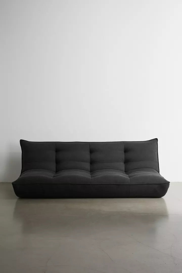 The Greta XL Sleeper Sofa, featuring a sleek and modern black design, sits elegantly against a plain white wall on a polished concrete floor. This sofa boasts a low profile with plush cushioning and a slightly curved backrest for added comfort.