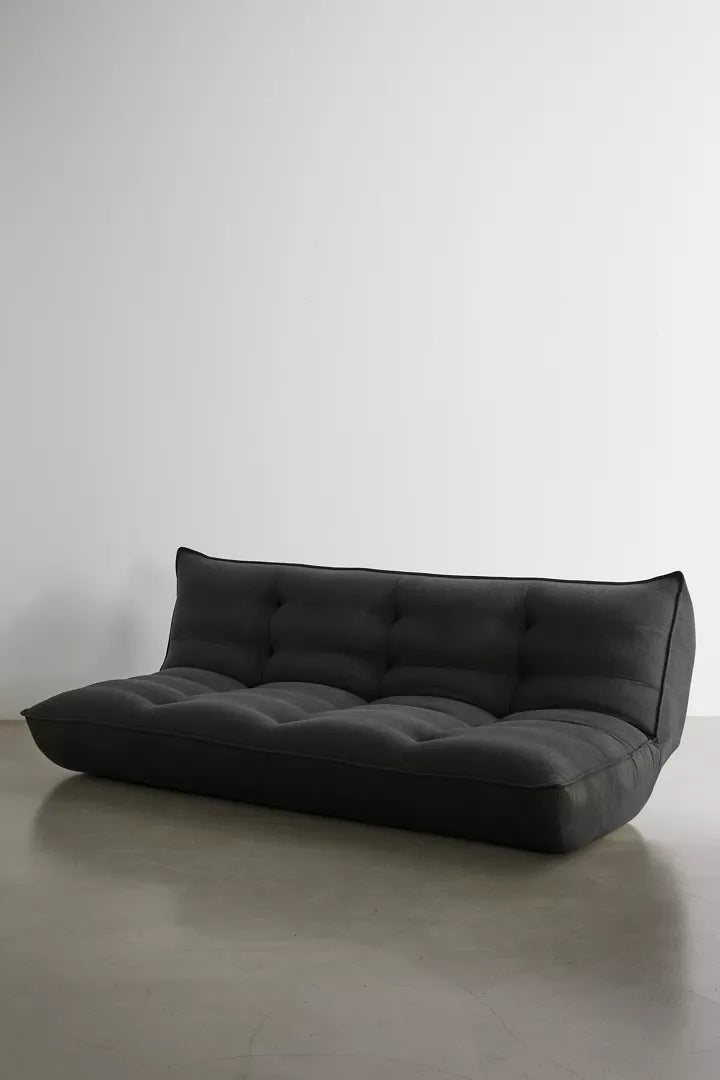 The Greta XL Sleeper Sofa, with its modern black tufted design, sits against a plain white wall on a polished concrete floor. Its minimalist style features a low back and no armrests, offering a sleek and contemporary appearance.