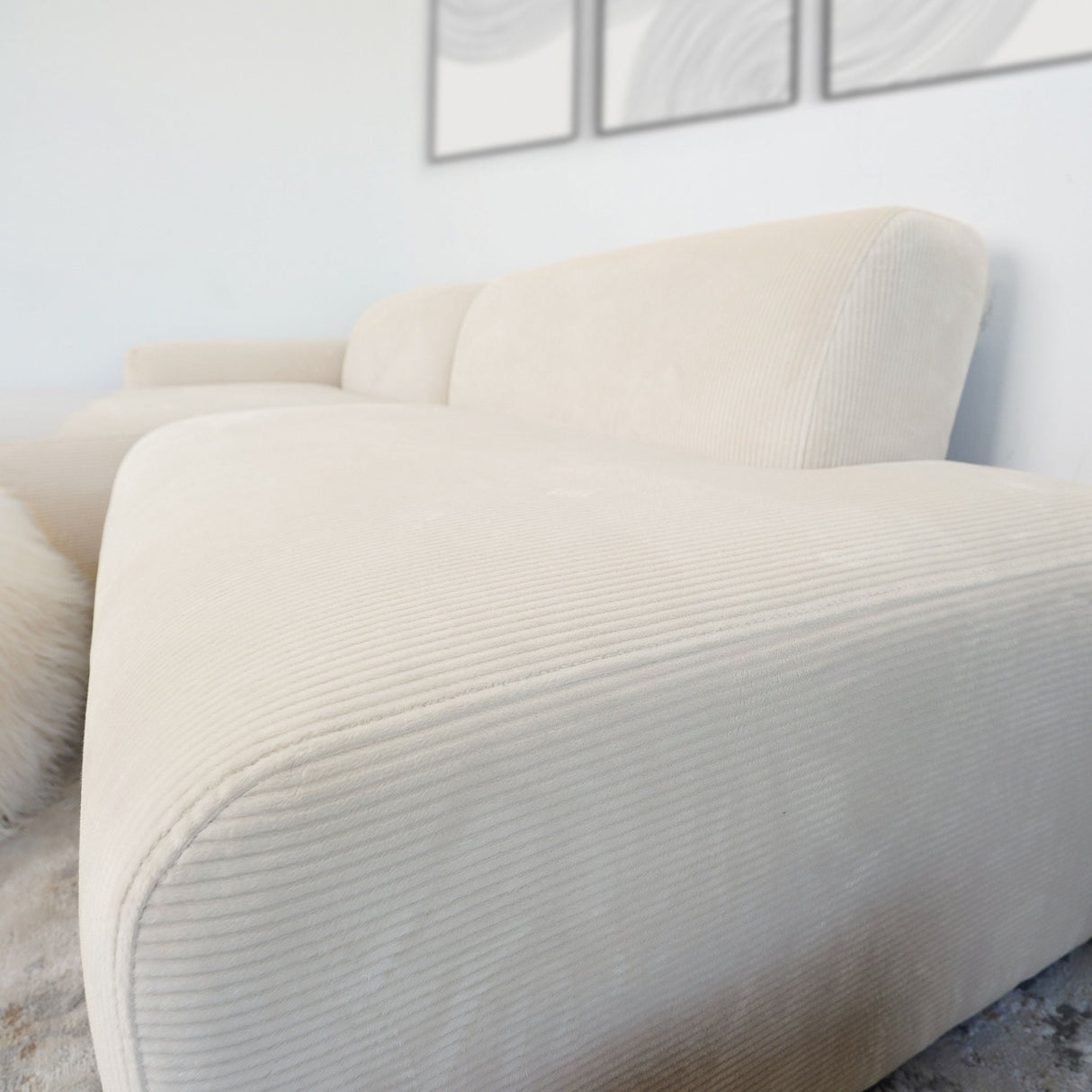 The Mar High-Quality Sectional Sofa, showcasing a sleek beige design with ribbed texture, stands out against a light wall adorned with abstract art. The carpeted flooring enhances the cozy ambiance of the minimalist space.