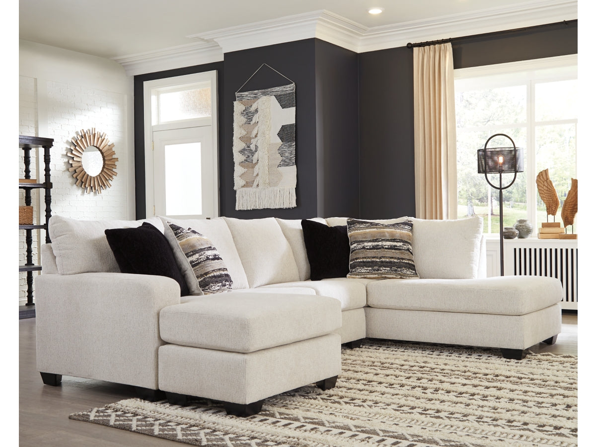 A modern living room showcasing the Cambri 2-Piece Sectional with Chaise in light beige, elegantly adorned with a mix of patterned and solid pillows. The space features a dark accent wall highlighted by abstract art, a large window dressed with curtains, and a textured rug underfoot. Minimalist decor adds the finishing touch to this serene setting.
