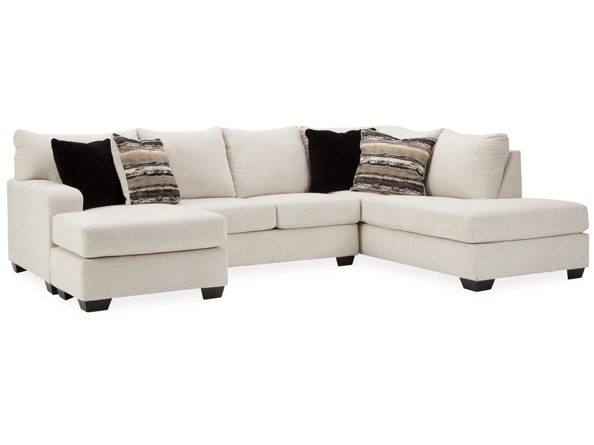 The Cambri 2-Piece Sectional with Chaise features a large beige design with a left chaise and right arm, complemented by three black pillows and two patterned pillows, all set against a white backdrop.