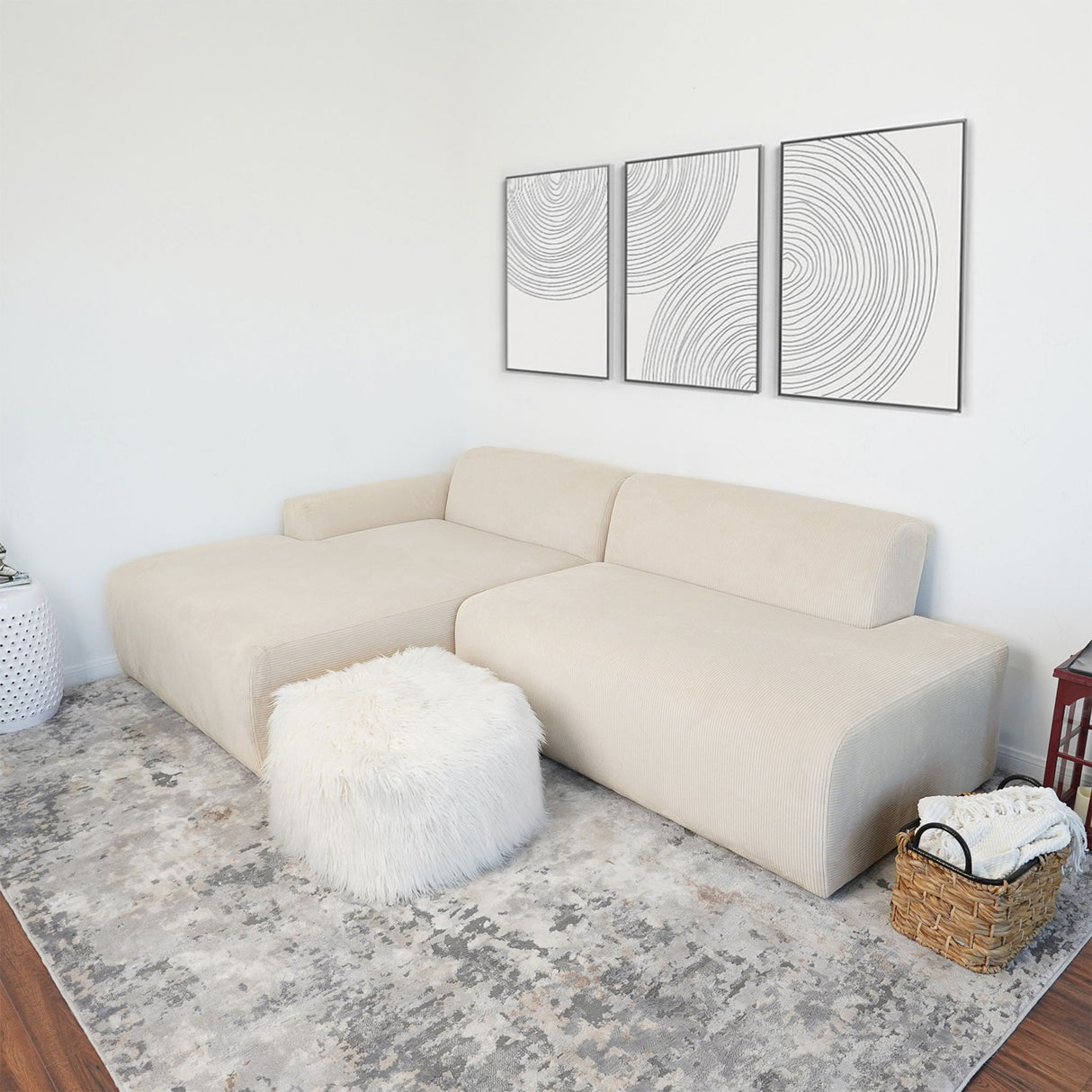A minimalist living room features the Mar - High-Quality Sectional Sofa in beige, a fluffy white ottoman, and a gray patterned rug. Three abstract line art pieces adorn the wall, while a woven basket and white planter add decorative touches.